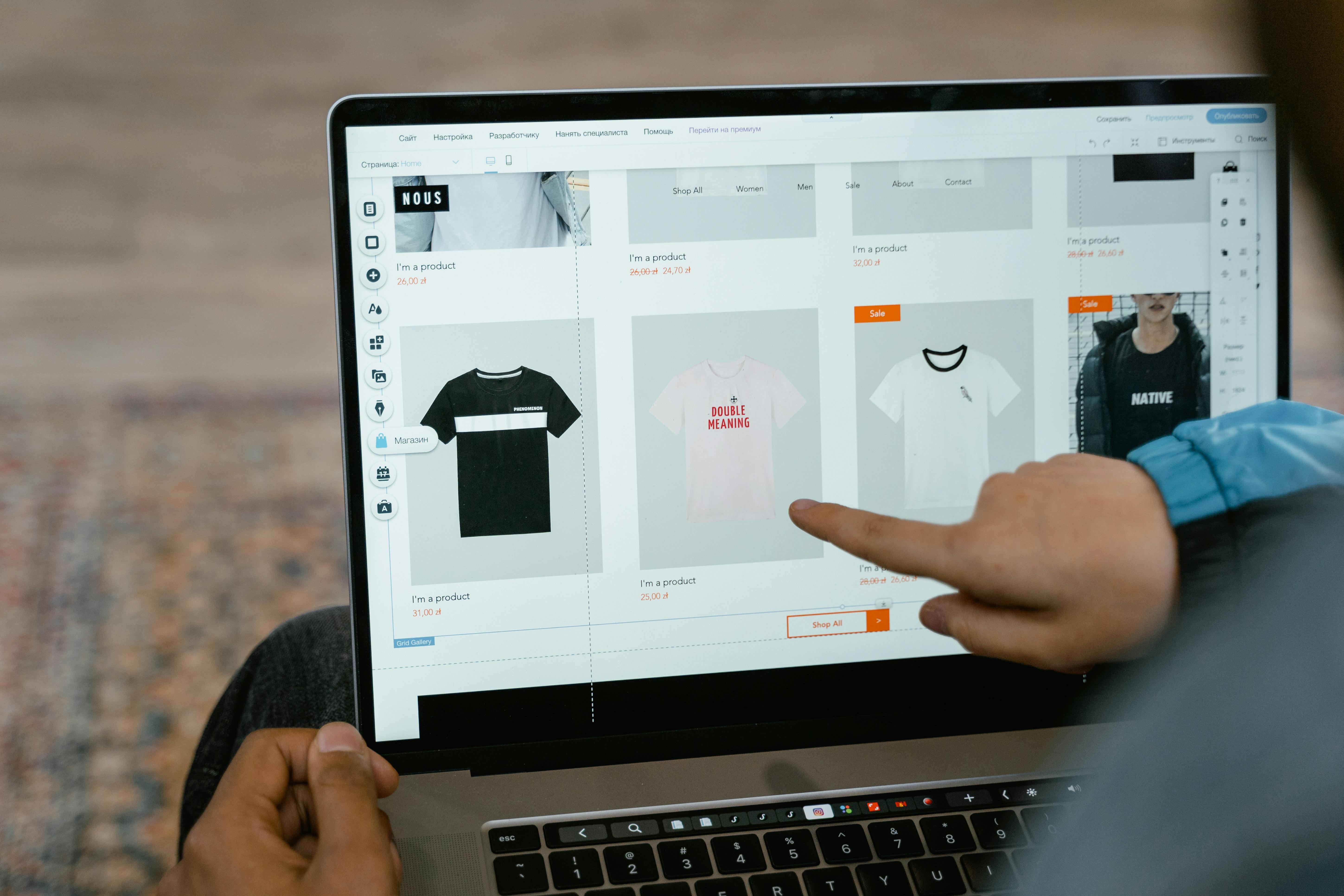 Person using a laptop to edit an online clothing store; t-shirt product cards are displayed on the screen with prices and sale tags, while a hand points at the UI on the right.