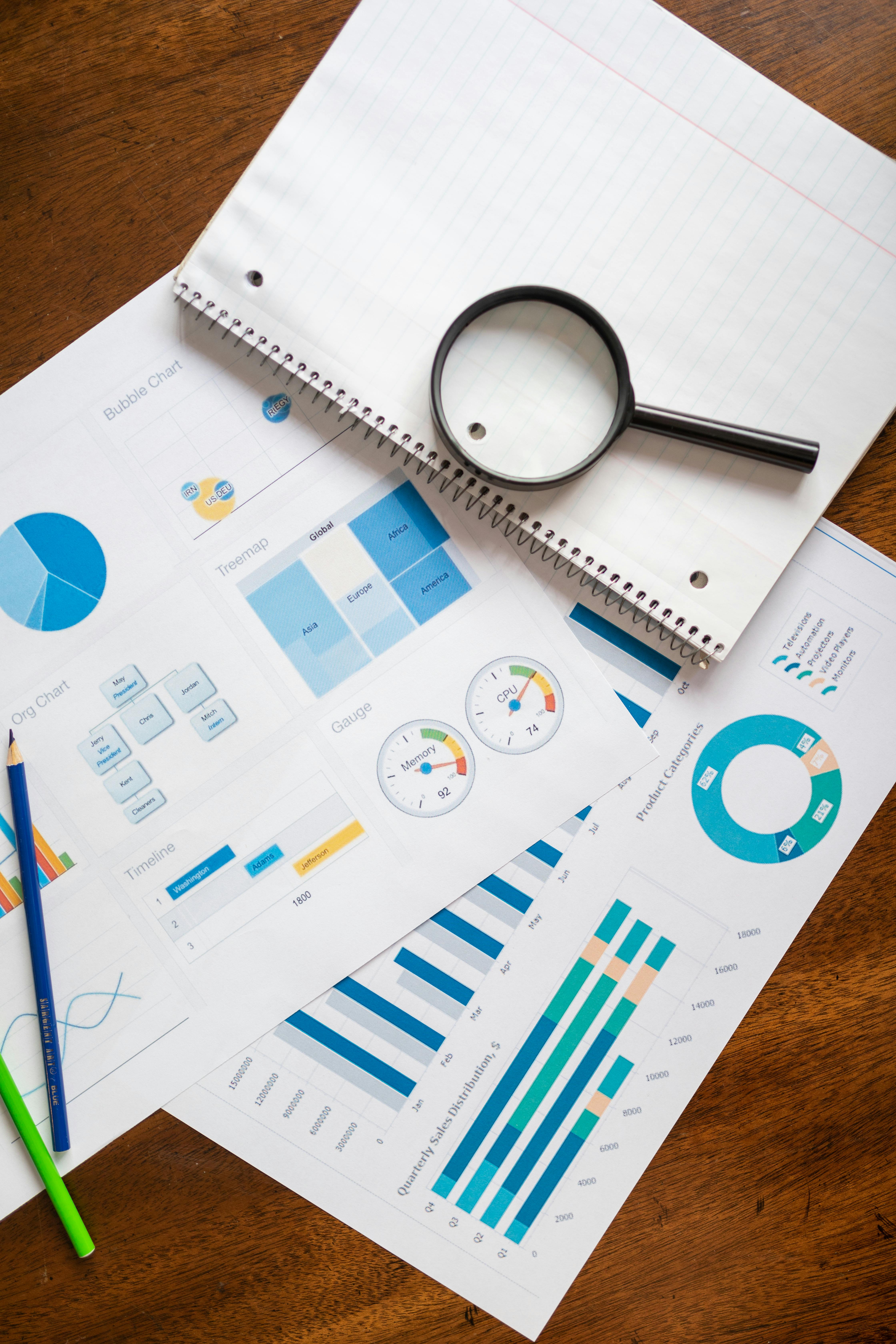 Open pages of business reports with blue charts and graphs on a wooden desk; a magnifying glass rests atop the documents.