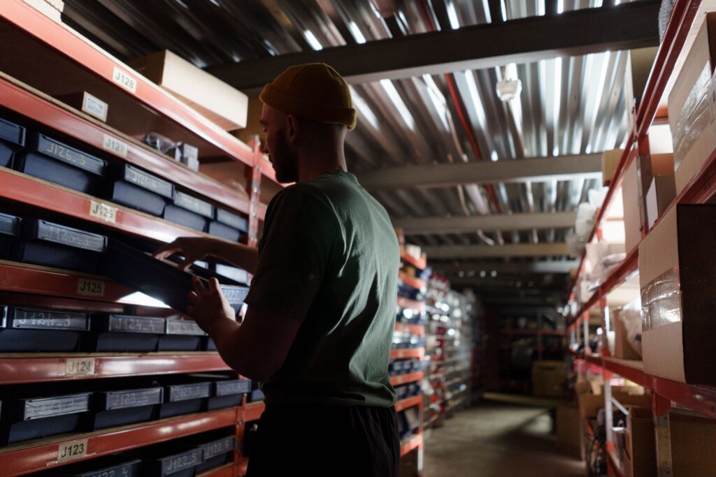 Man wearing a mustard beanie selecting a labeled bin from orange warehouse shelves, viewed in profile