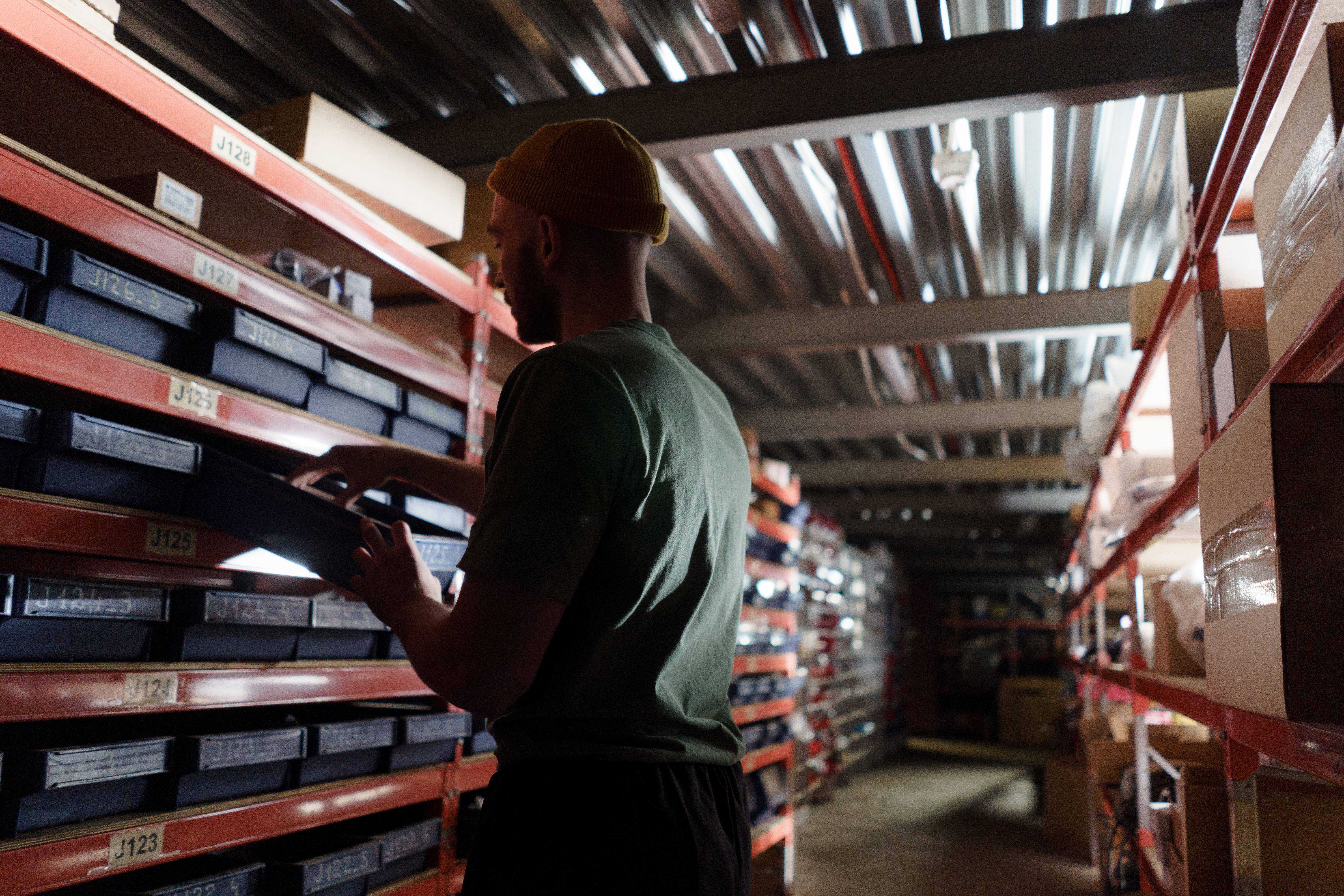 Man wearing a mustard beanie selecting a labeled bin from orange warehouse shelves, viewed in profile