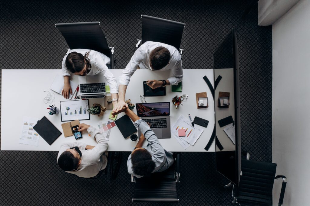 Overhead view of a team meeting around a white table with laptops, charts and papers scattered nearby.