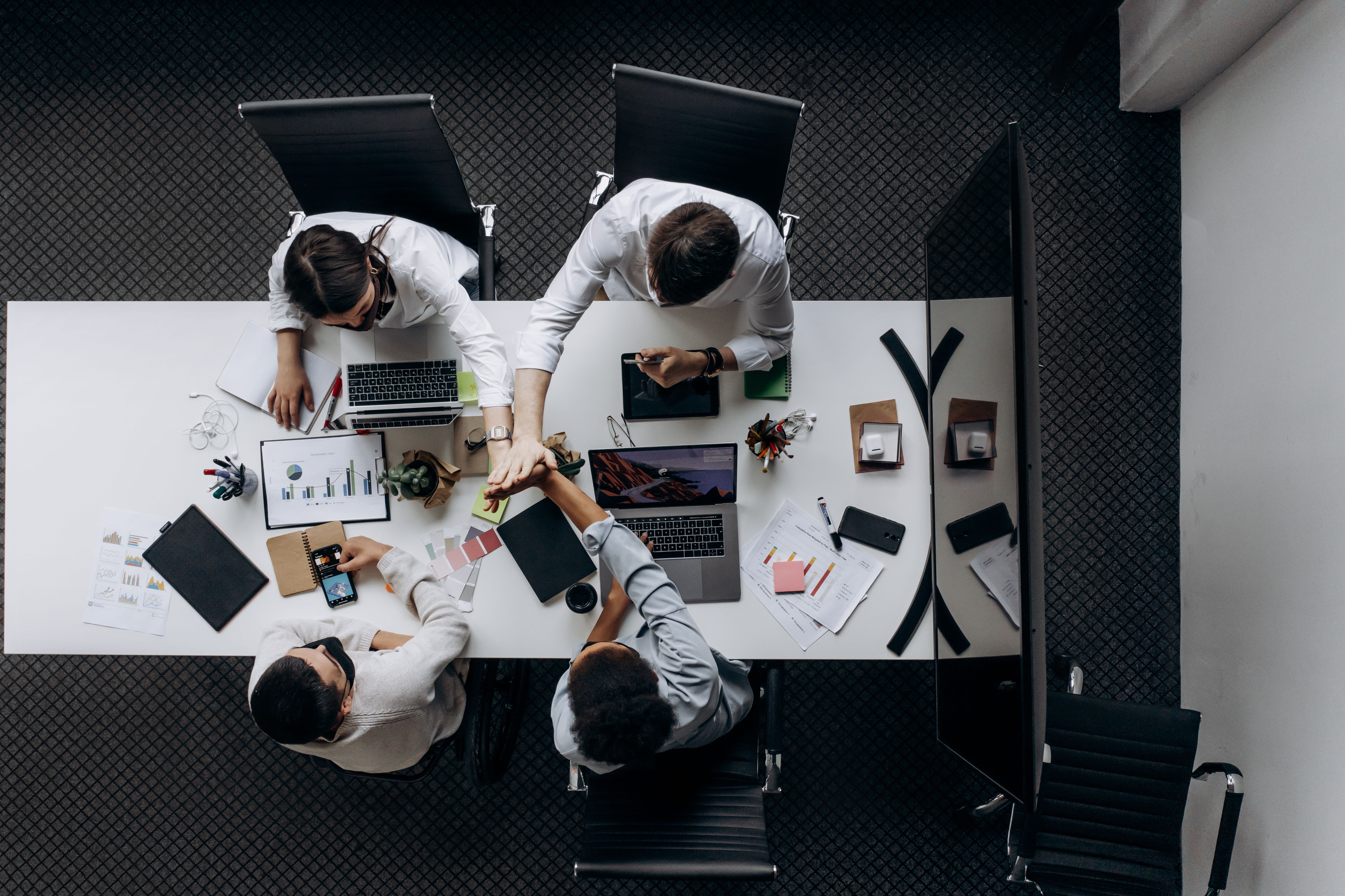 Overhead view of a team meeting around a white table with laptops, charts and papers scattered nearby.